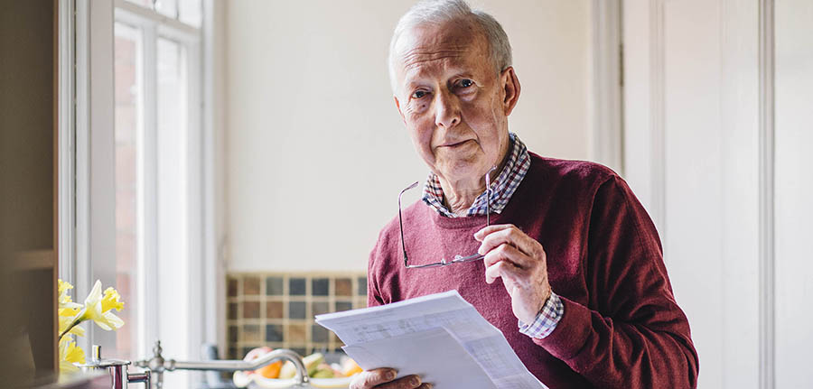 Senior man is standing in the kitchen of his home with bills in his hand, looking at the camera with a worried expression.